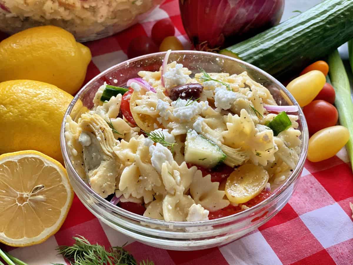 A glass bowl of bow tie pasta salad on a red and white checkered tablecloth.