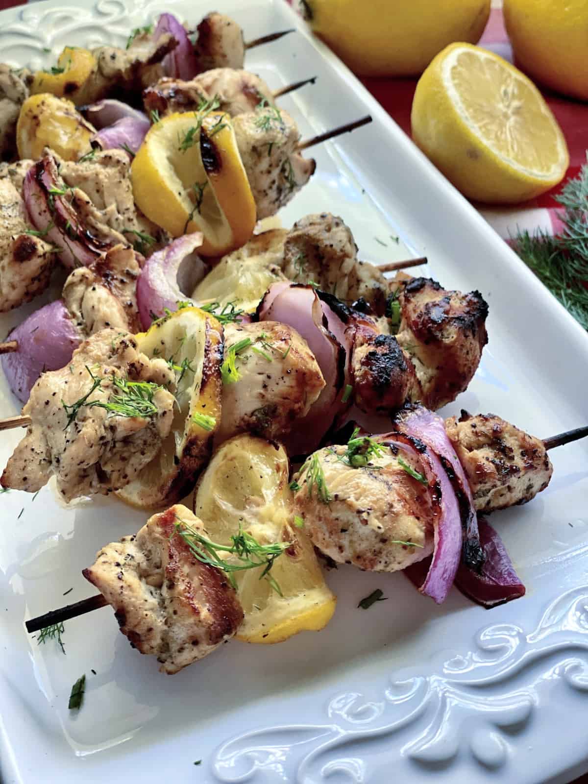 Chicken, lemon, and red onion on wooden skewers resting on a white platter. 