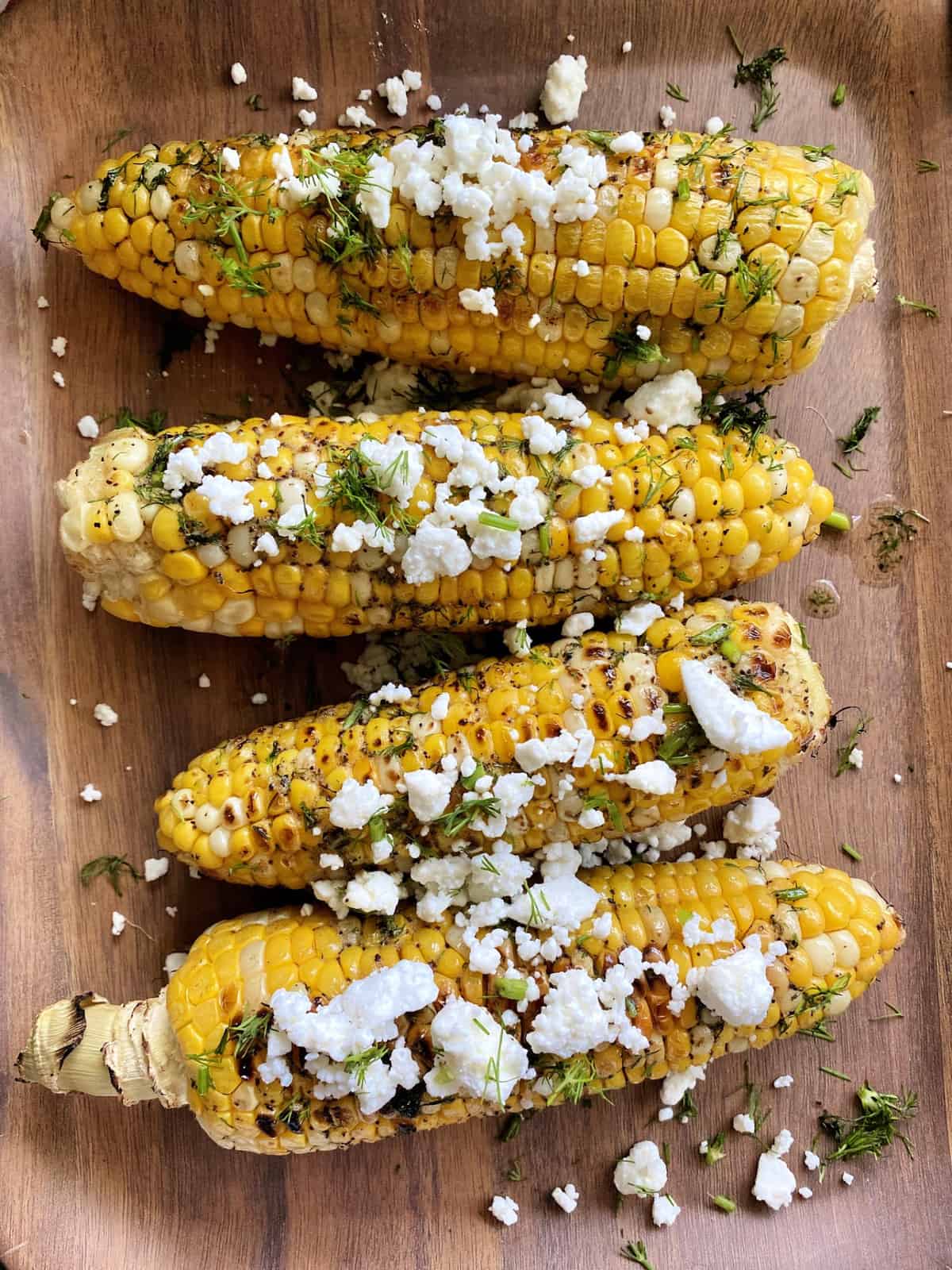 Top view of corn on the cob on a wooden plate topped with feta and fresh dill.