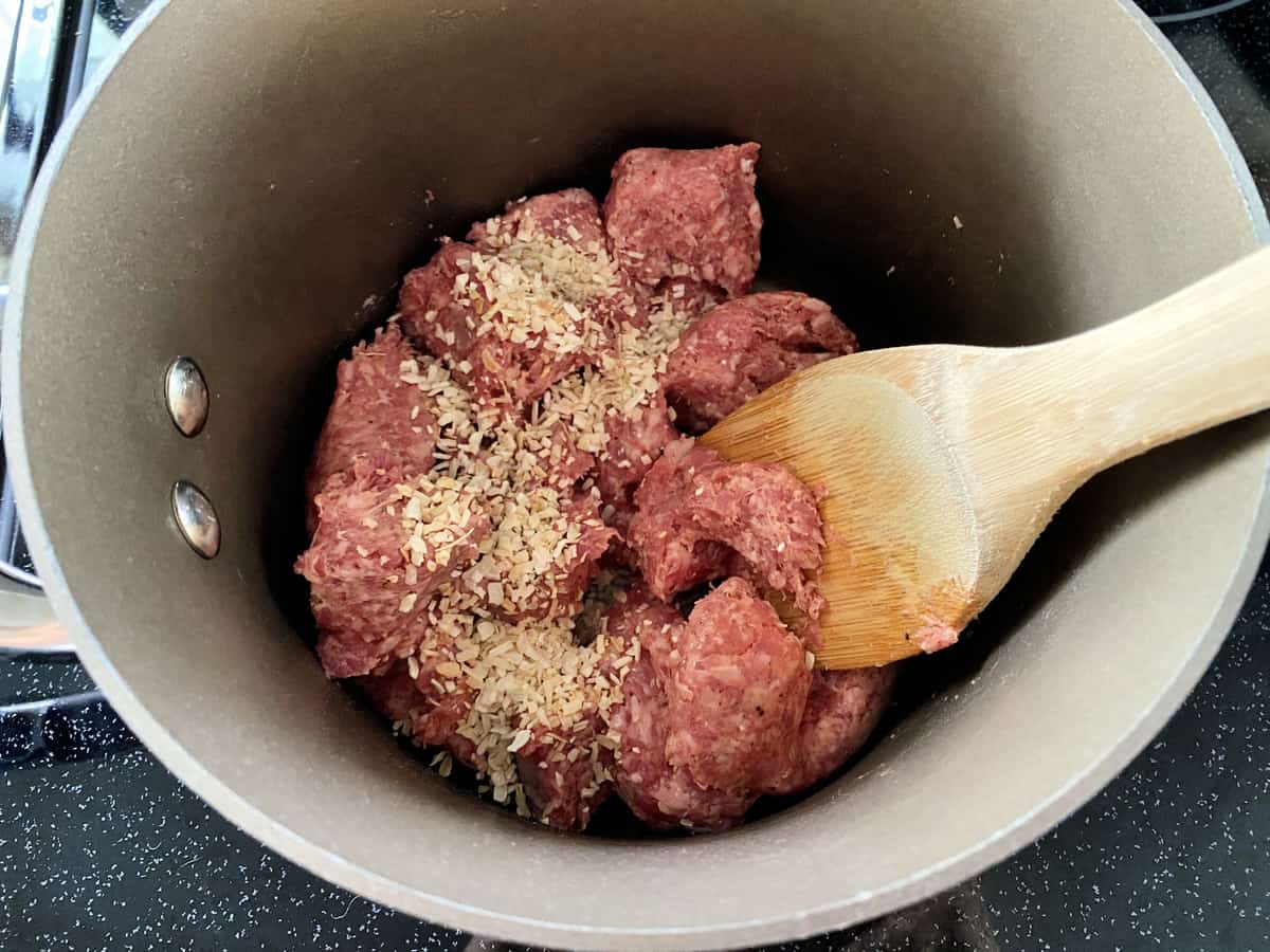 Sauce pan with ground sausage and dried onion flakes with a wooden spoon.