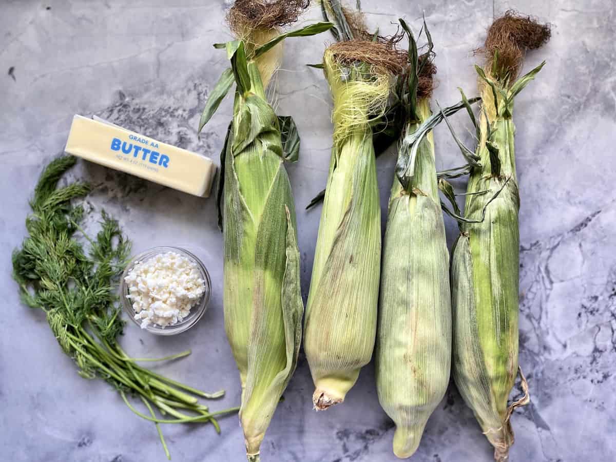 Corn with husk on, salted butter, bowl of crumbled feta, and fresh dill.