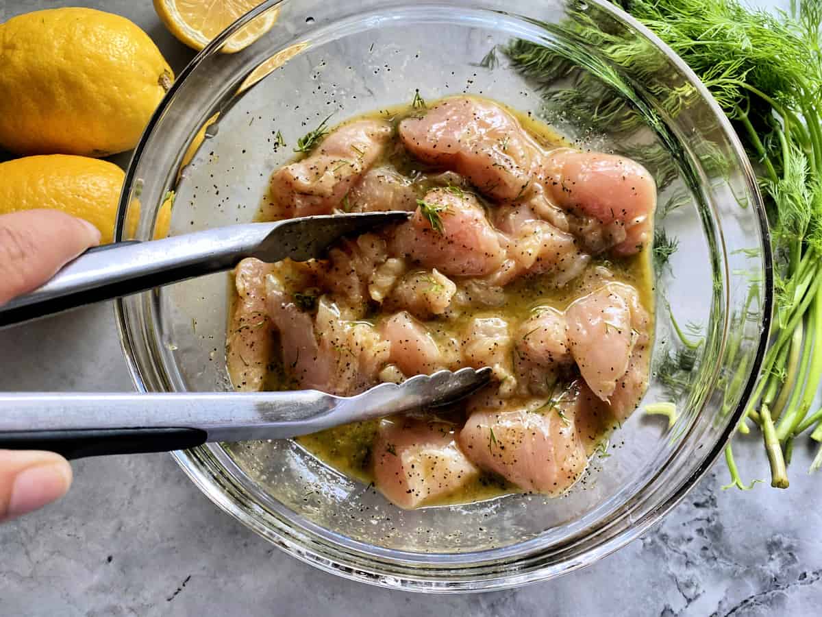 Raw chicken in a glass bowl of Greek marinade with tongs tossing chicken. 