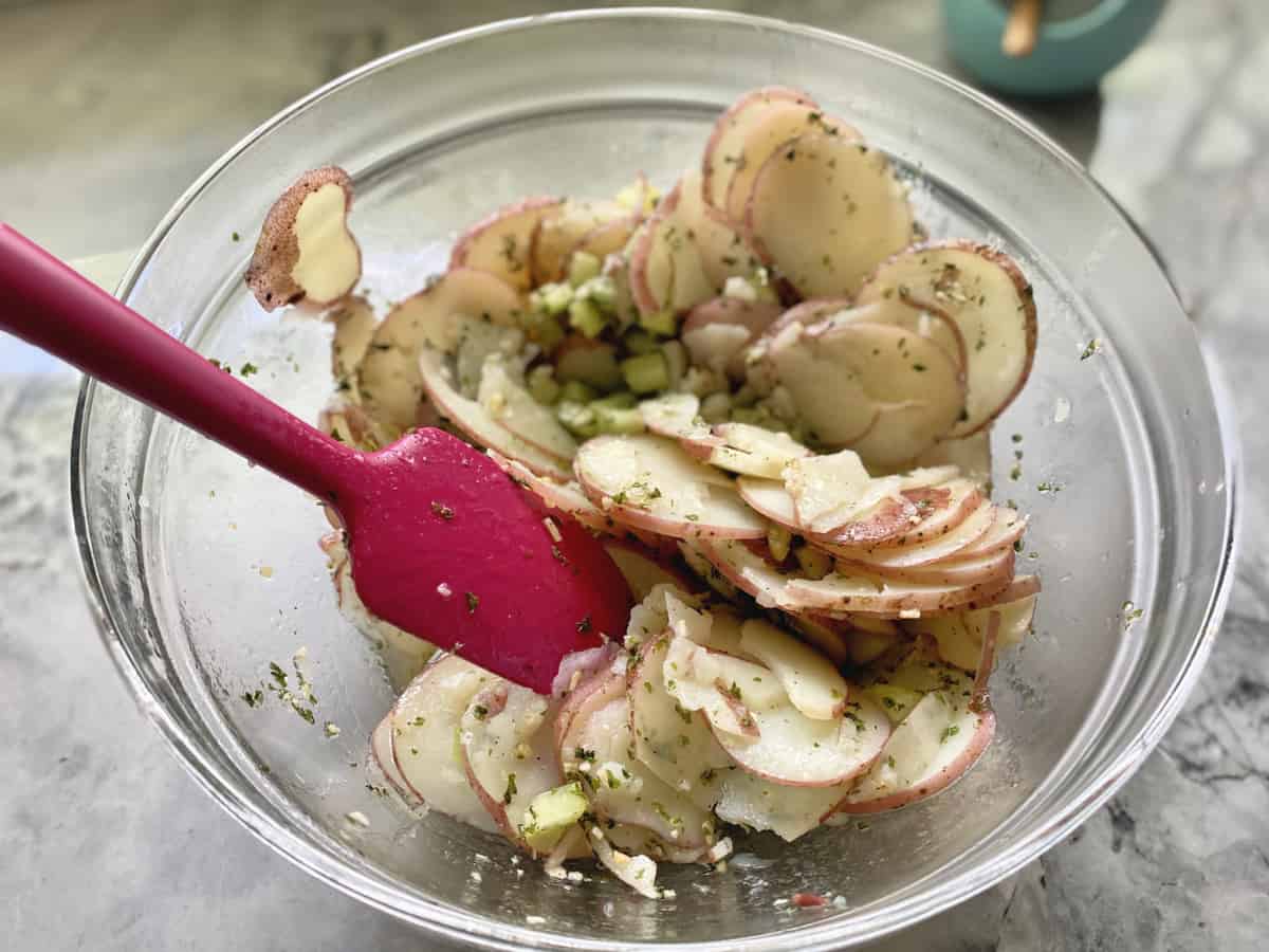 Top view of a glass bowl with sliced potatoes mixed in dressing with a pink spoon.
