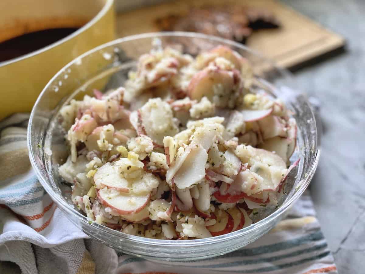 Thinly sliced cooked red potatoes with dressing in a glass bowl sitting on a dishcloth.