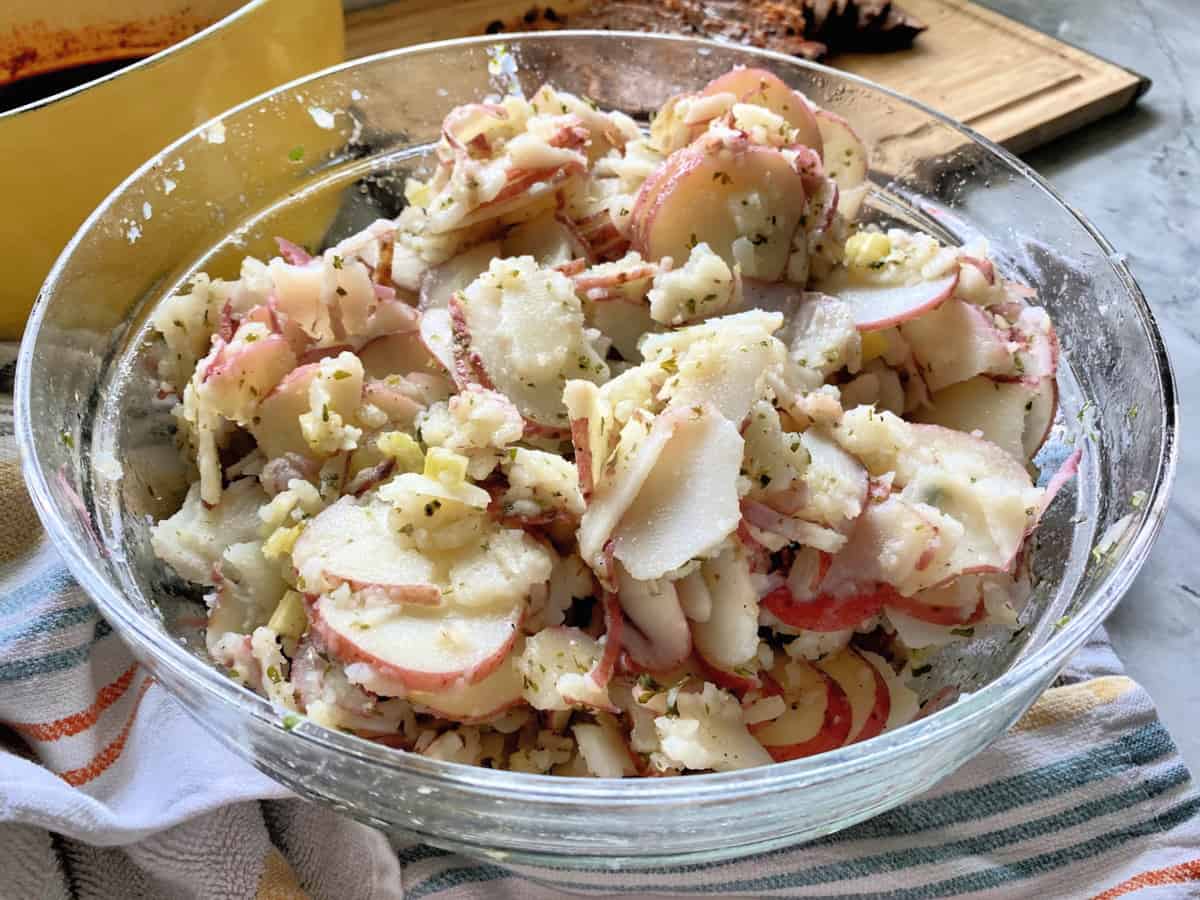 Glass bowl sitting on a dishtowel with sliced potatoes and dressing in it.