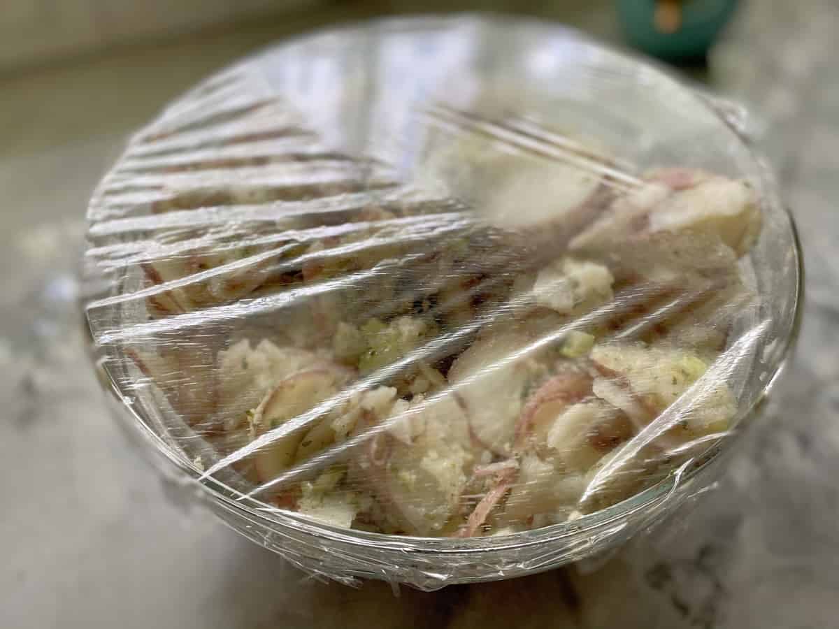 Glass bowl with potato salad in it wrapped with plastic wrap on counter.