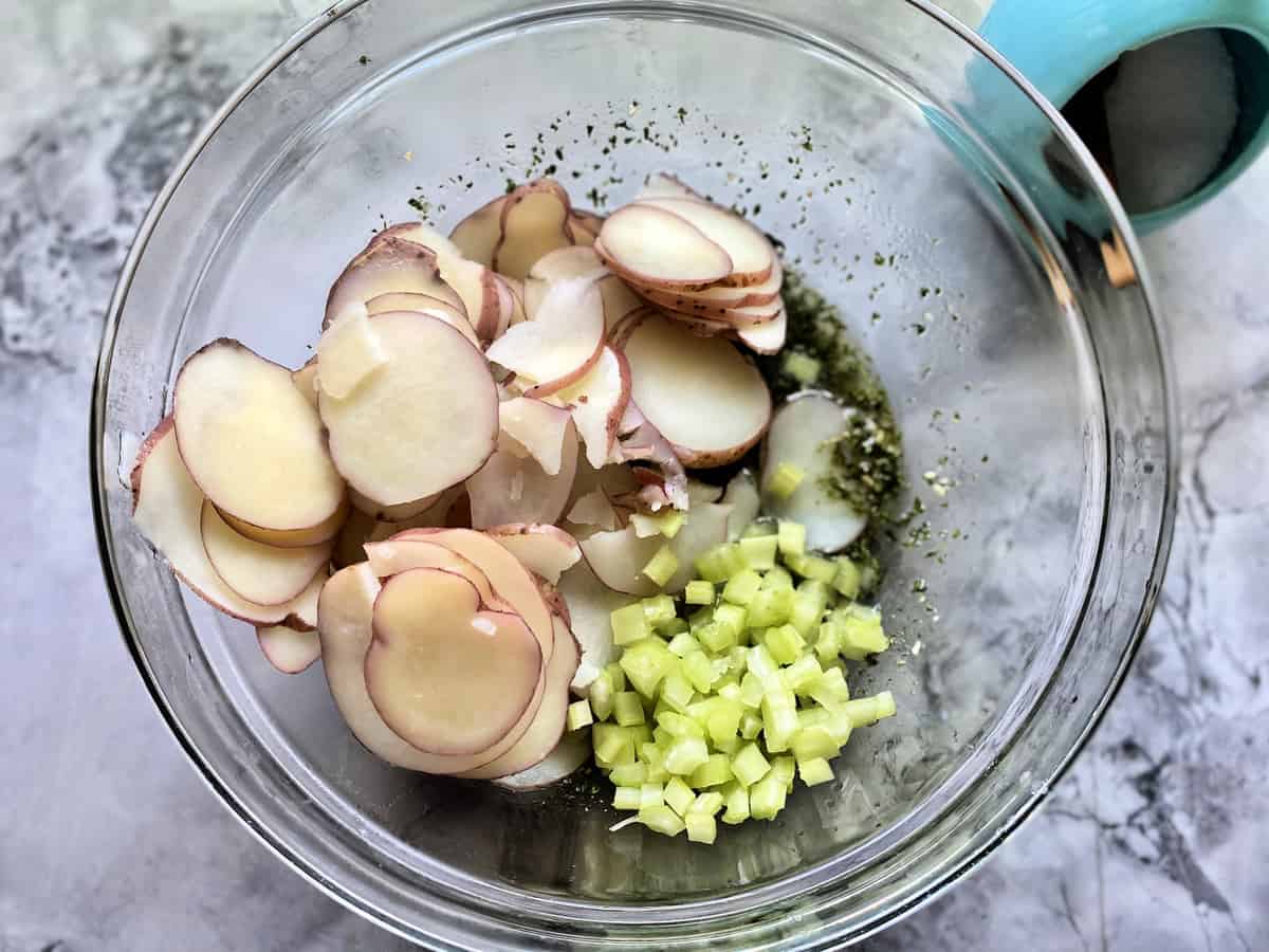 Top view of a glass bowl with sliced cooked potatoes, celery, and dressing. 