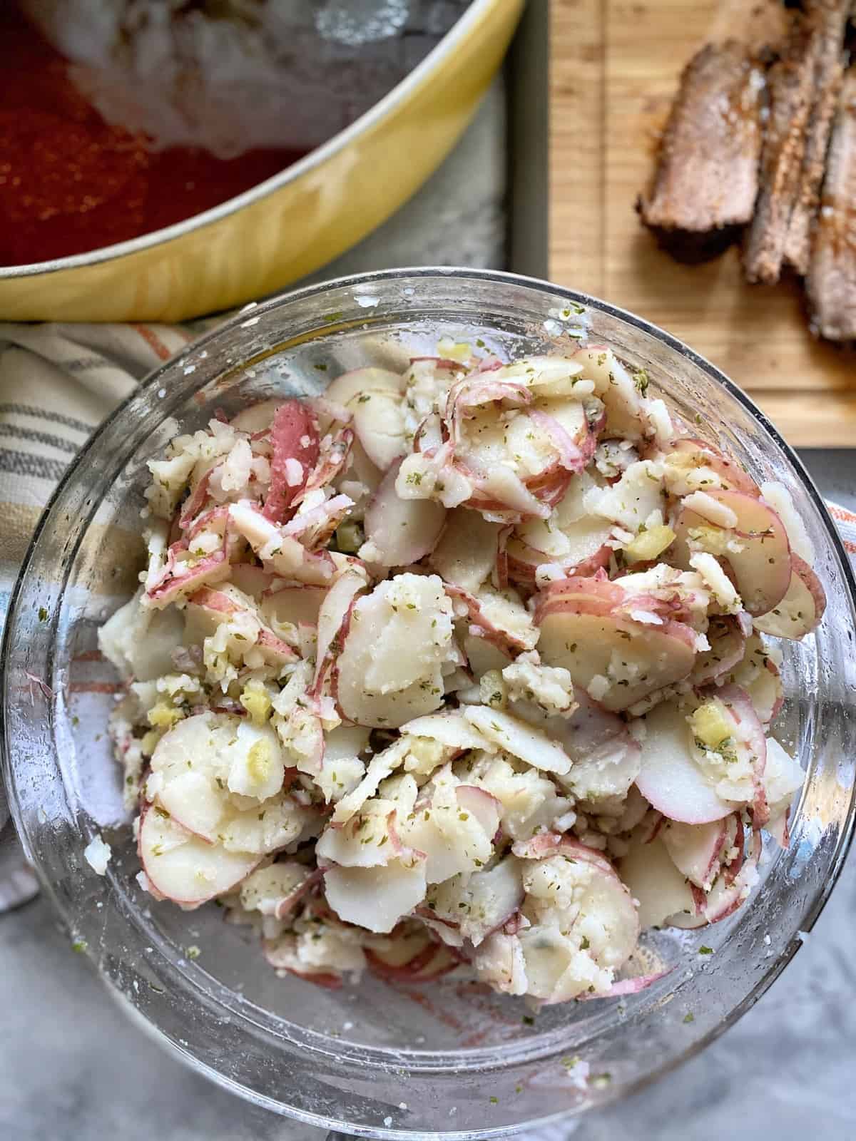 Top view of mixed potato salad in a glass bowl with brisket and sauce in the background. 