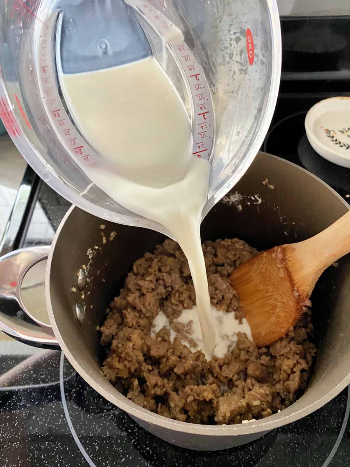 A measuring cup pouring milk into a sauce pan of cooked ground pork.