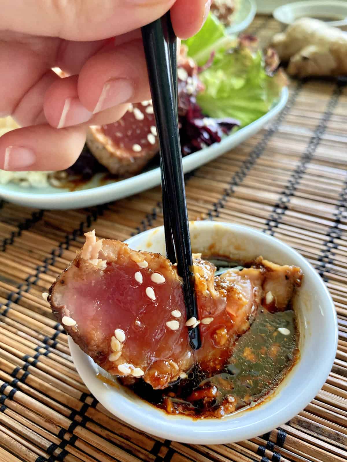 Female hand holding hopsticks dipping tuna into a small white sauce dish. 