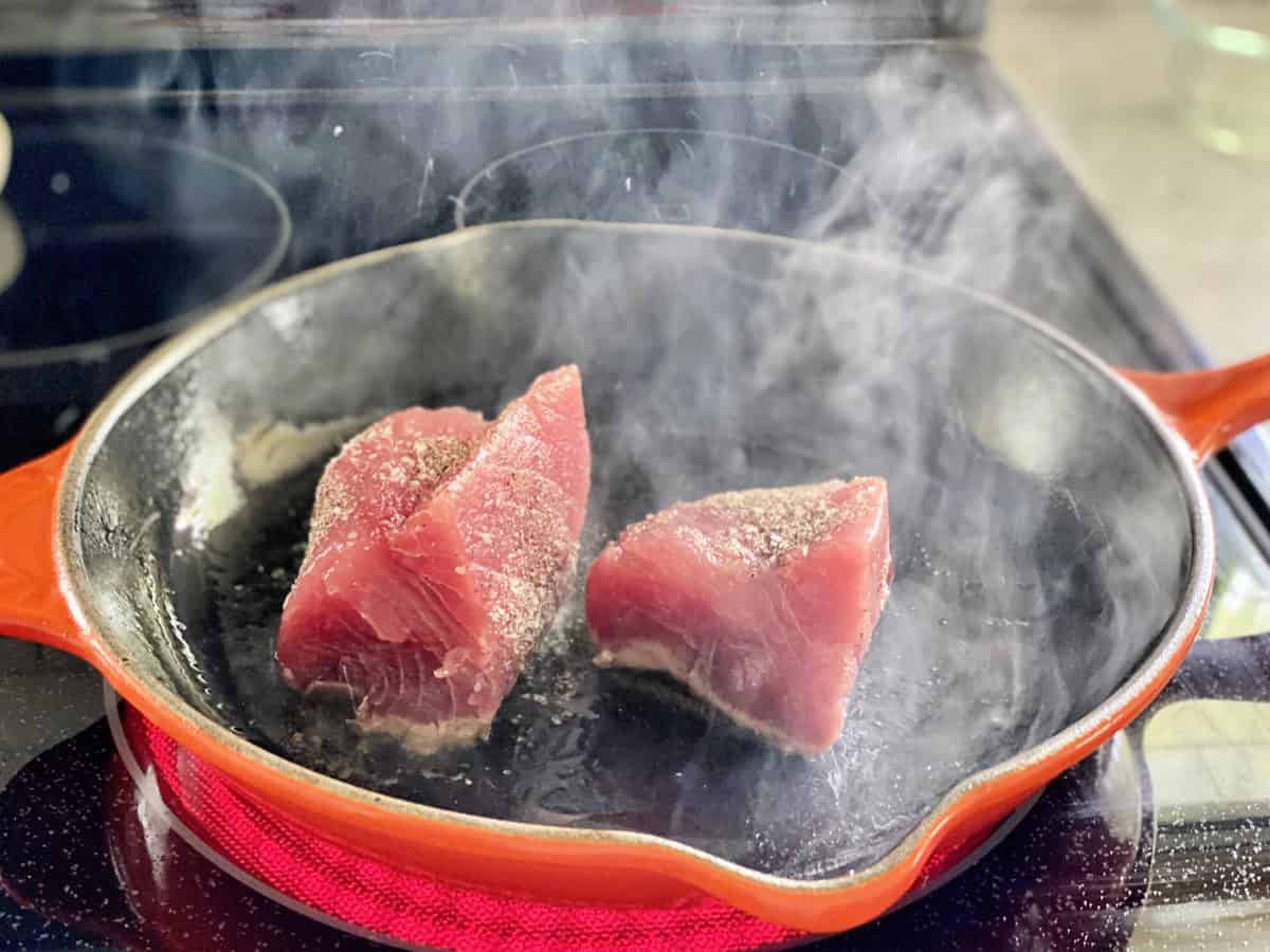 Orange cast iron skillet on a stove top searing tuna with smoke rising from pan.