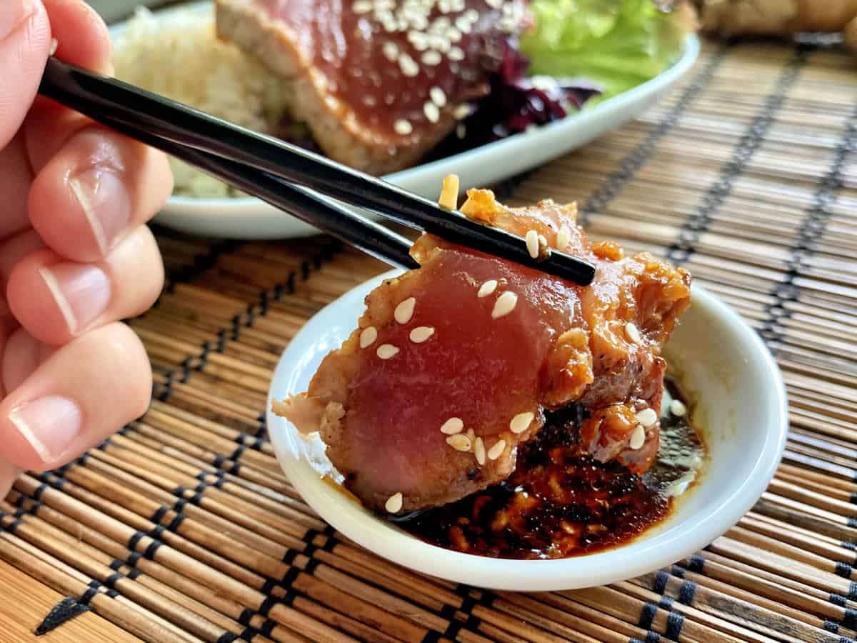 Female hand holding chopsticks with seared tuna dipped in a sauce with sesame seeds.