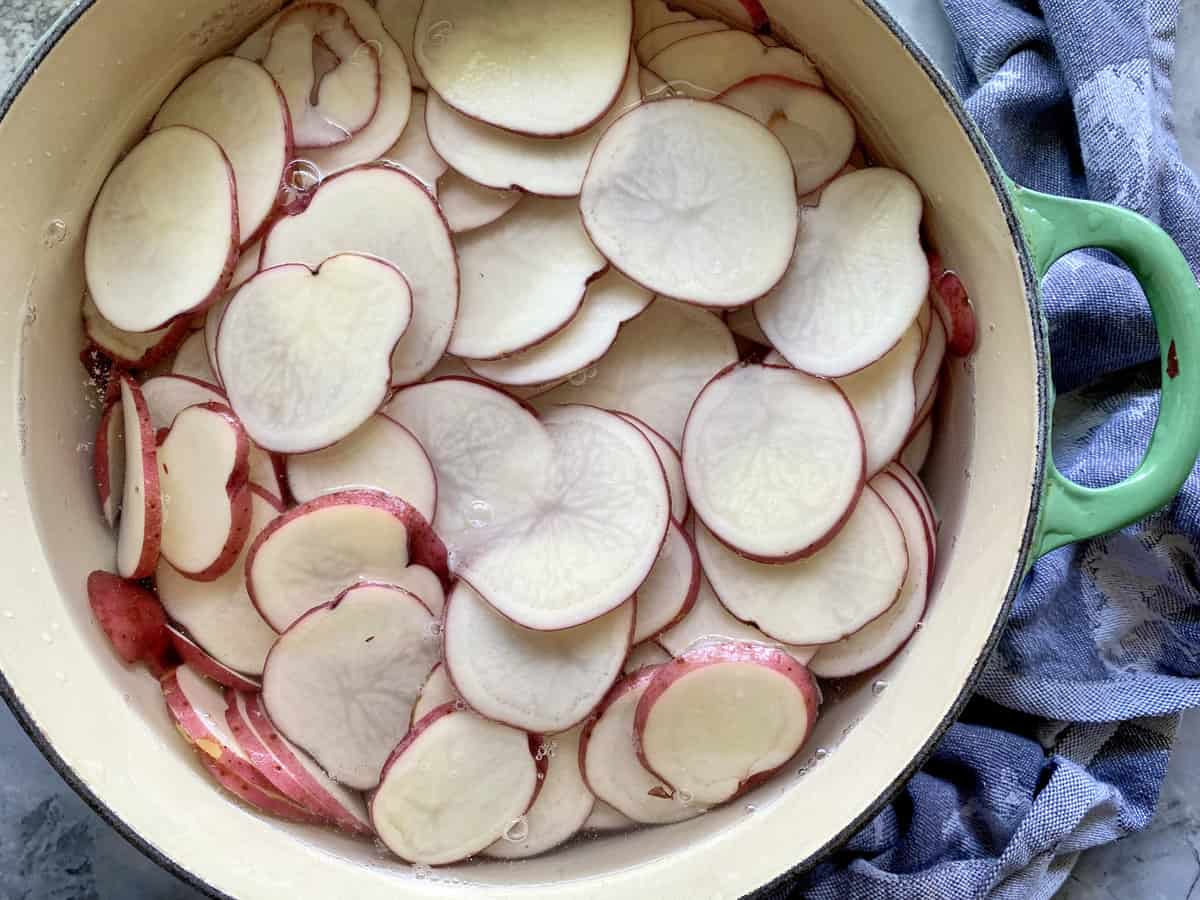 Top view of sliced red potatoes soaking in water in a green pot. 