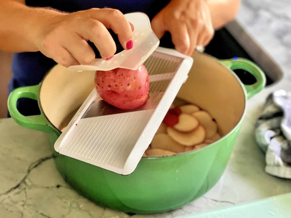 Pink painted nails slicing a red potato on a mandolin slicer over a green pot.