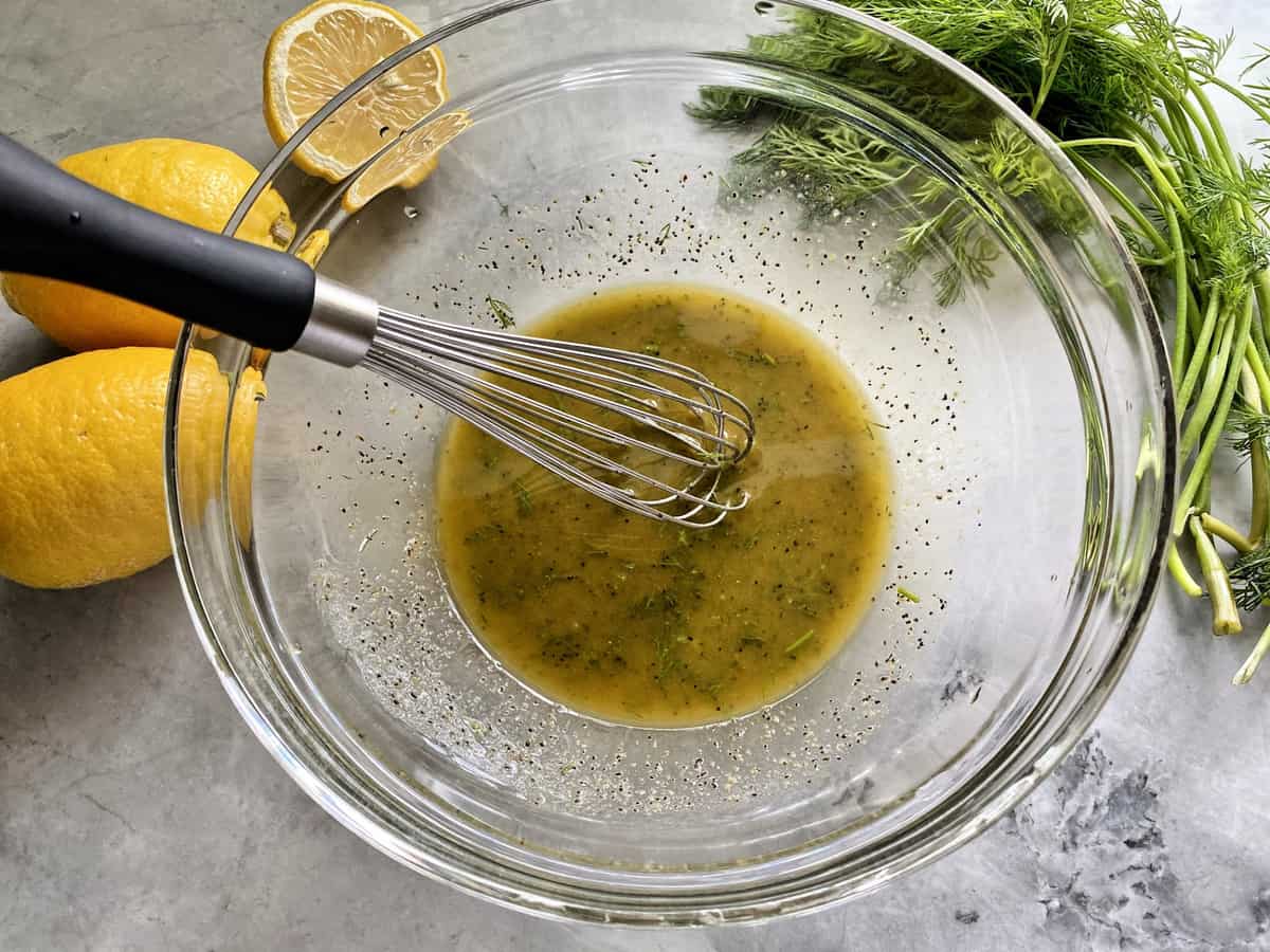 A glass bowl with homemade Greek dressing and whisk. Fresh dill and lemons surround the outside bowl.