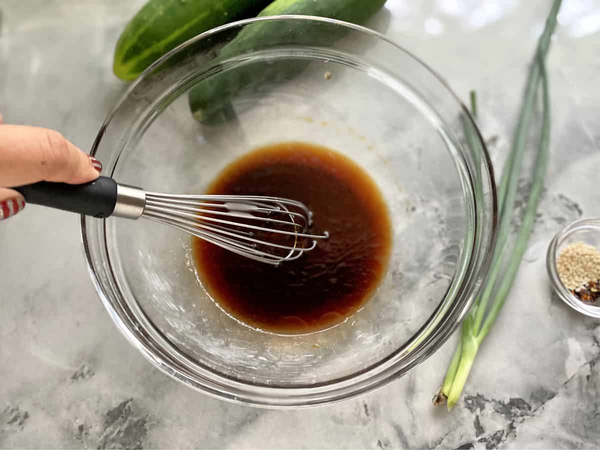 Hand with painted nails whisking brown dressing in a glass bowl on a marble countertop.