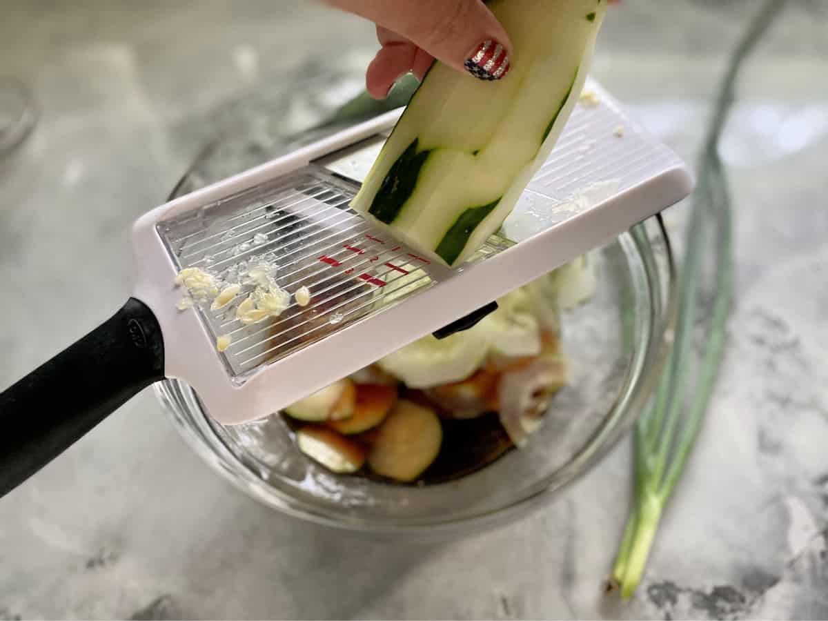 Hand with painted nails thinly slicing cucumbers on a mandolin slicer into a bowl.