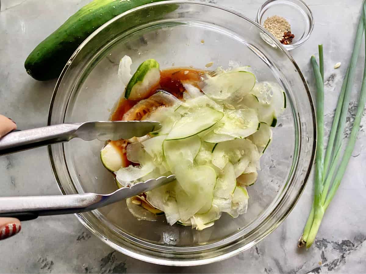 Tongs tossing thinly sliced cucumber in brown dressing in a glass bowl on a marble countertop.