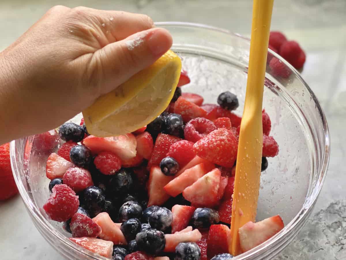 Hand squeezing lemon juice into a glass bowl of berries with orange spatula in bowl.