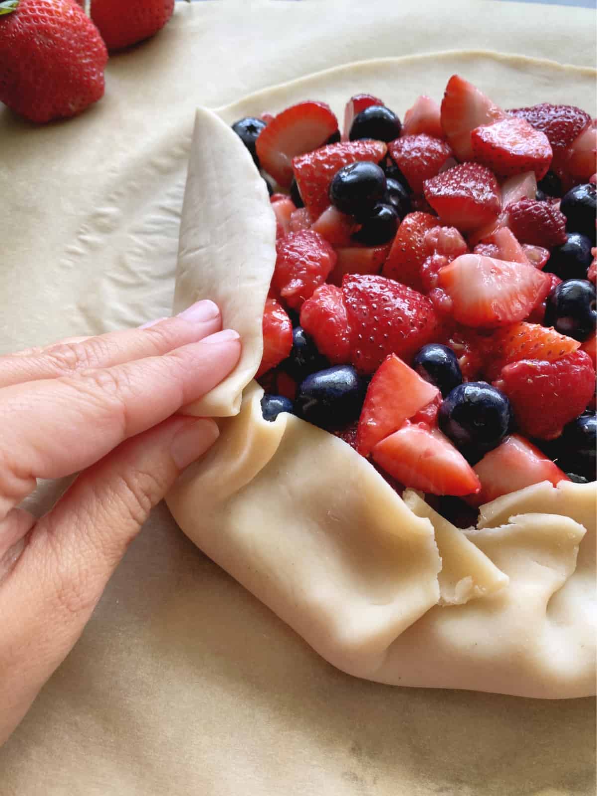 Hand folding pie crust around a pile of berries on brown parchment paper.