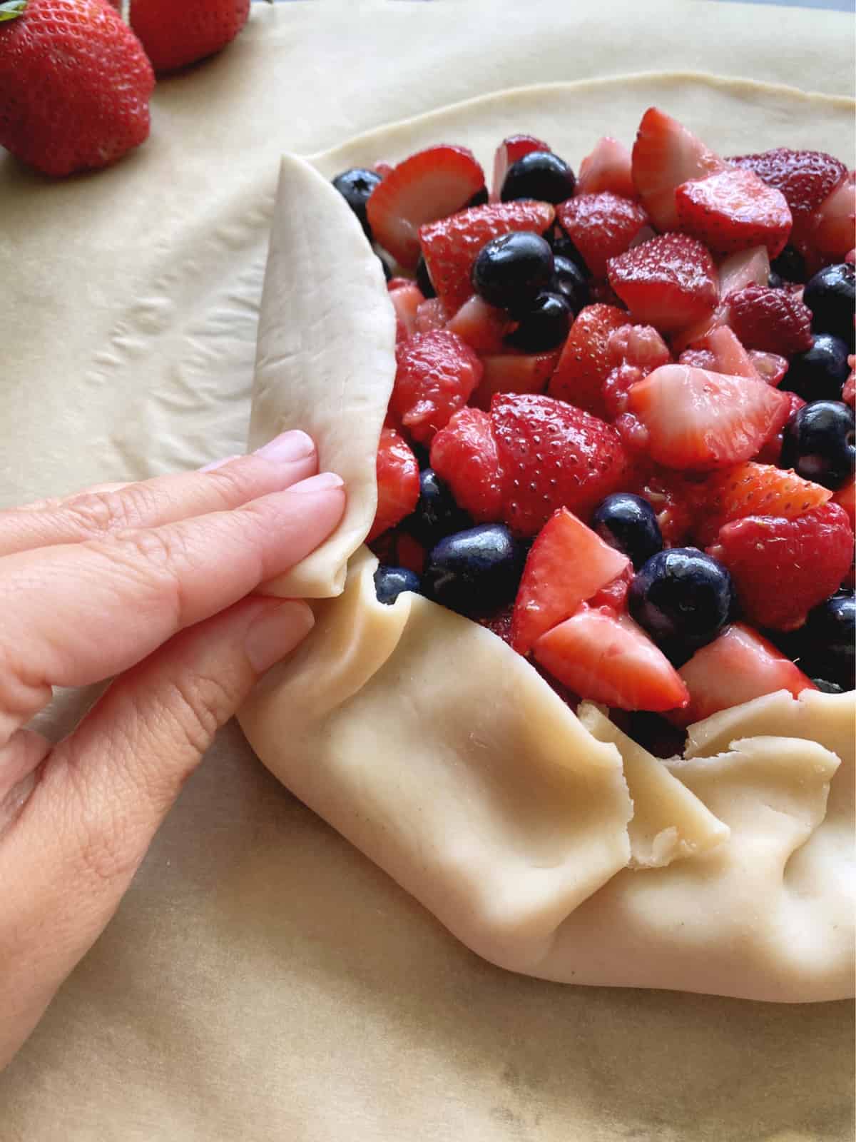 Hand folding pie crust around a pile of berries on brown parchment paper. 