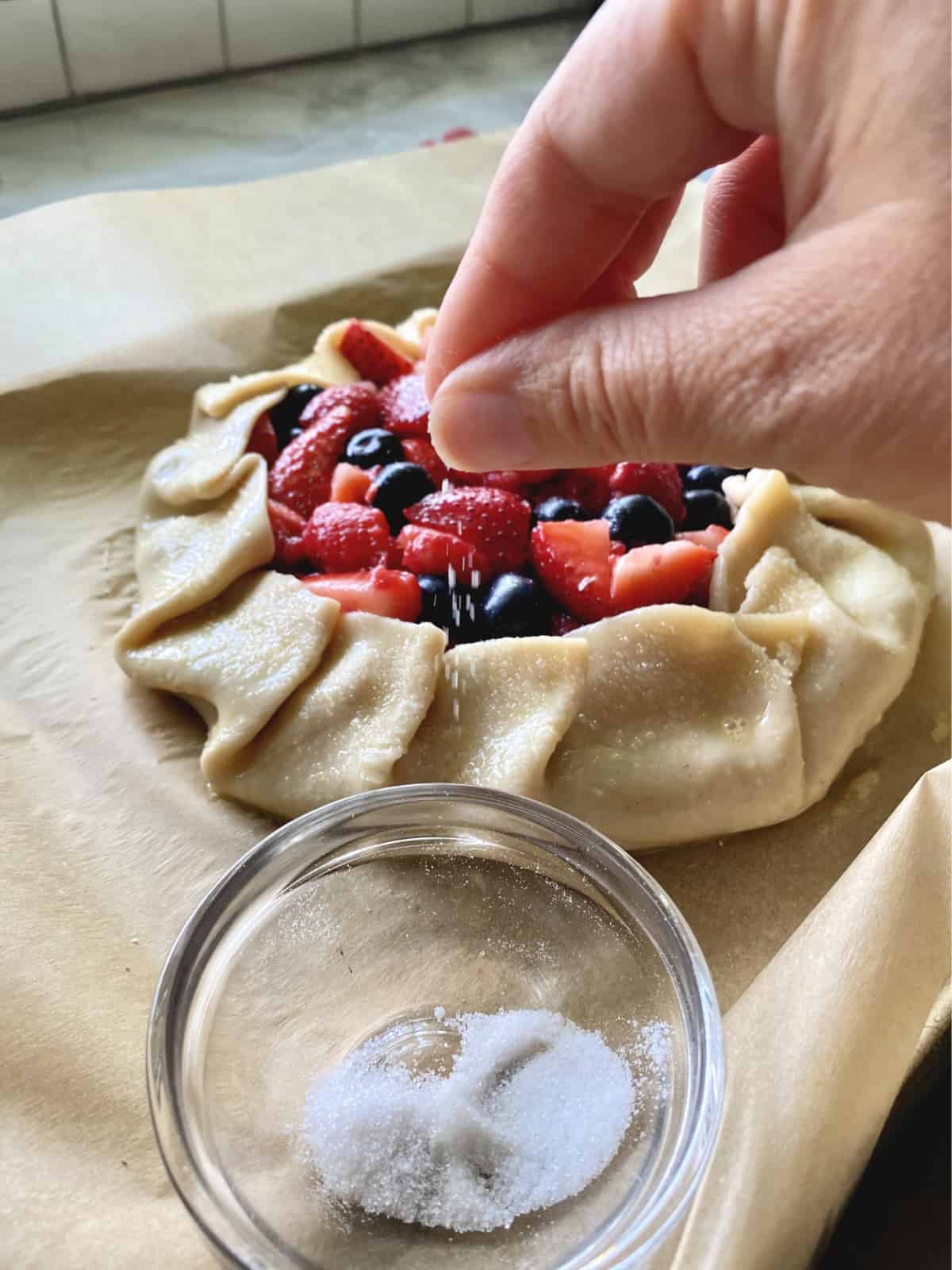 Hand sprinkling granulated sugar on an egg washed pie crust with berries in the center.