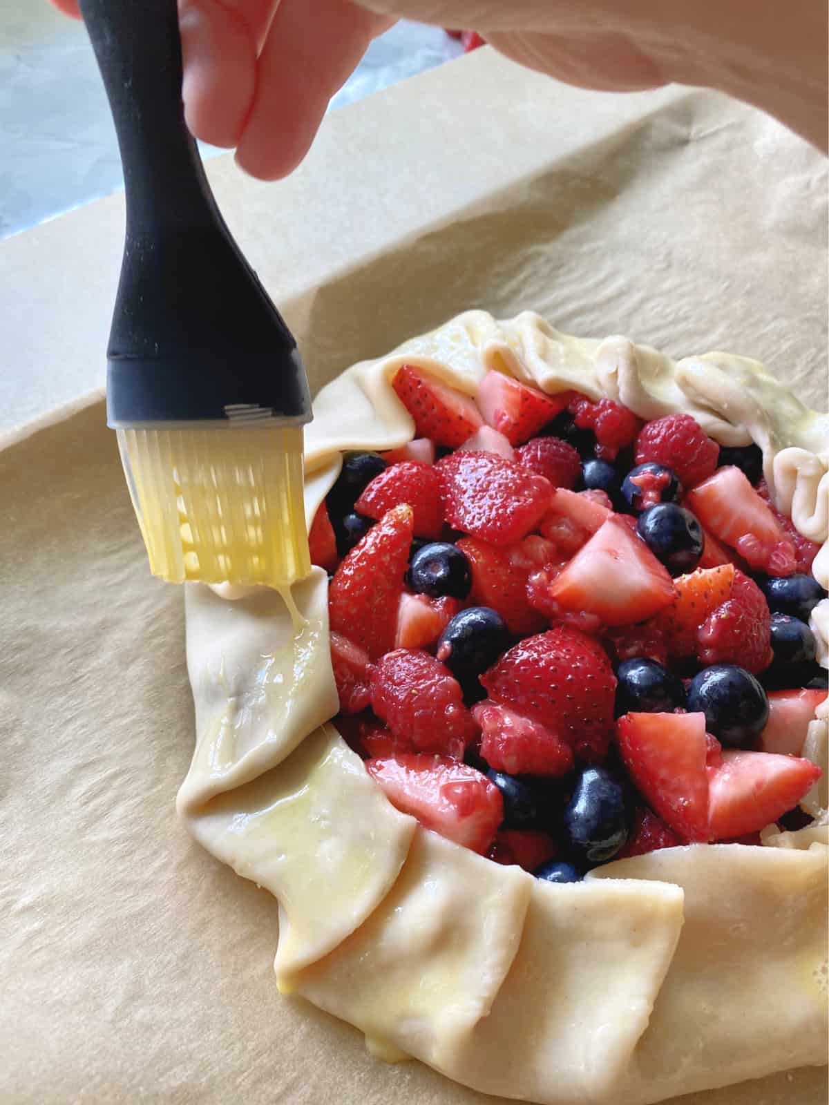 Hand holding silicone brush, brushing egg wash on the edge of pie crust. 