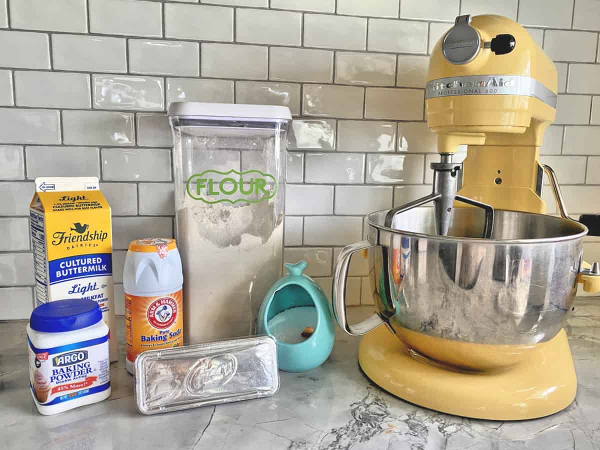 Buttermilk, baking powder, baking soda, flour, Crisco, salt next to yellow KitchenAid Stand Mixer.