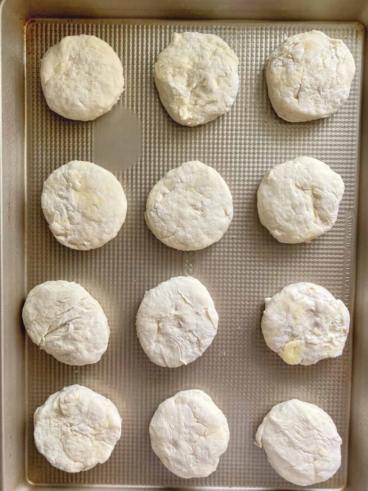 Top view of a dozen raw cut out biscuit dough on a baking sheet.