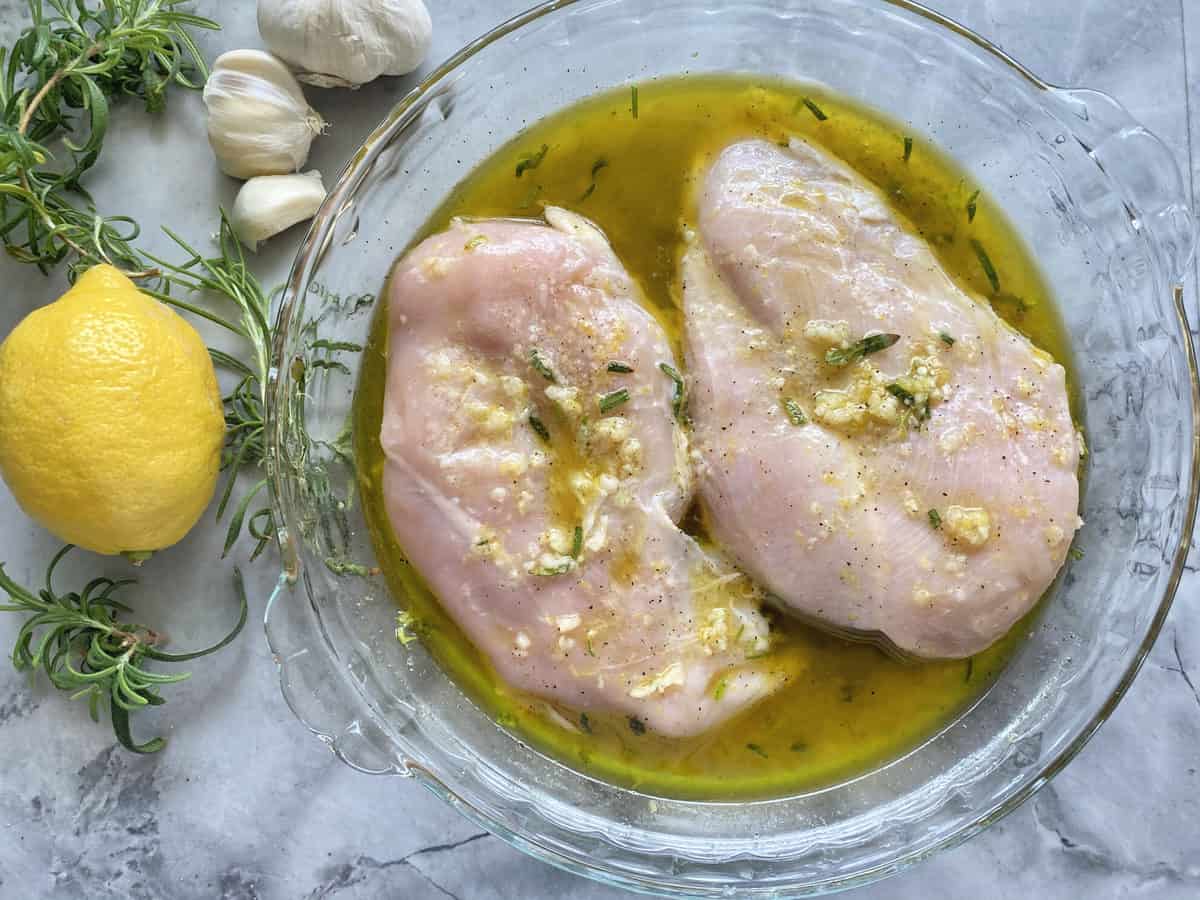 Top view of raw chicken breast in a garlic marinade with chopped rosemary leaves.