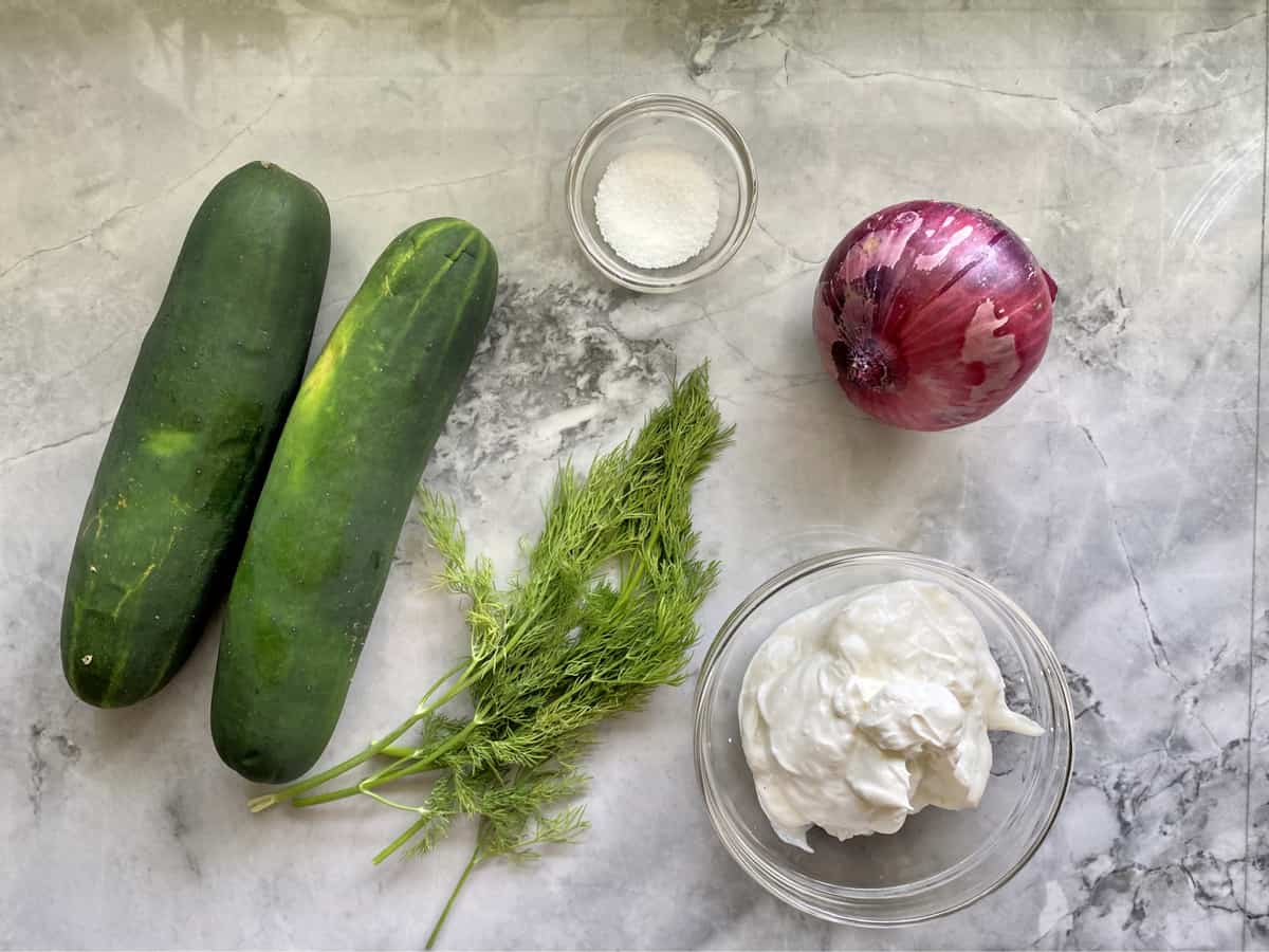 Ingredients on marble countertop: cucumber, fresh dill, salt, red onion, and sour cream.