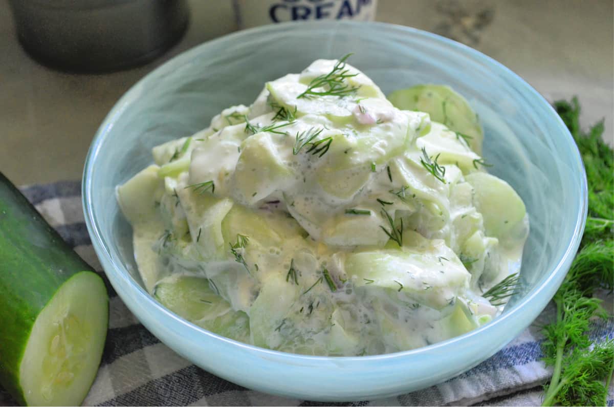 Blue bowl on a dish cloth with sliced cucumbers, white cream sauce, fresh dill, and onion.