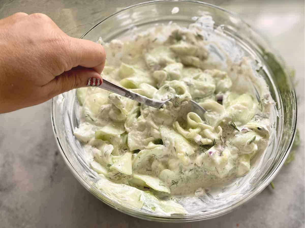 Hand mixing sliced cucumbers with red onion, sour cream, fresh dill in a glass bowl.