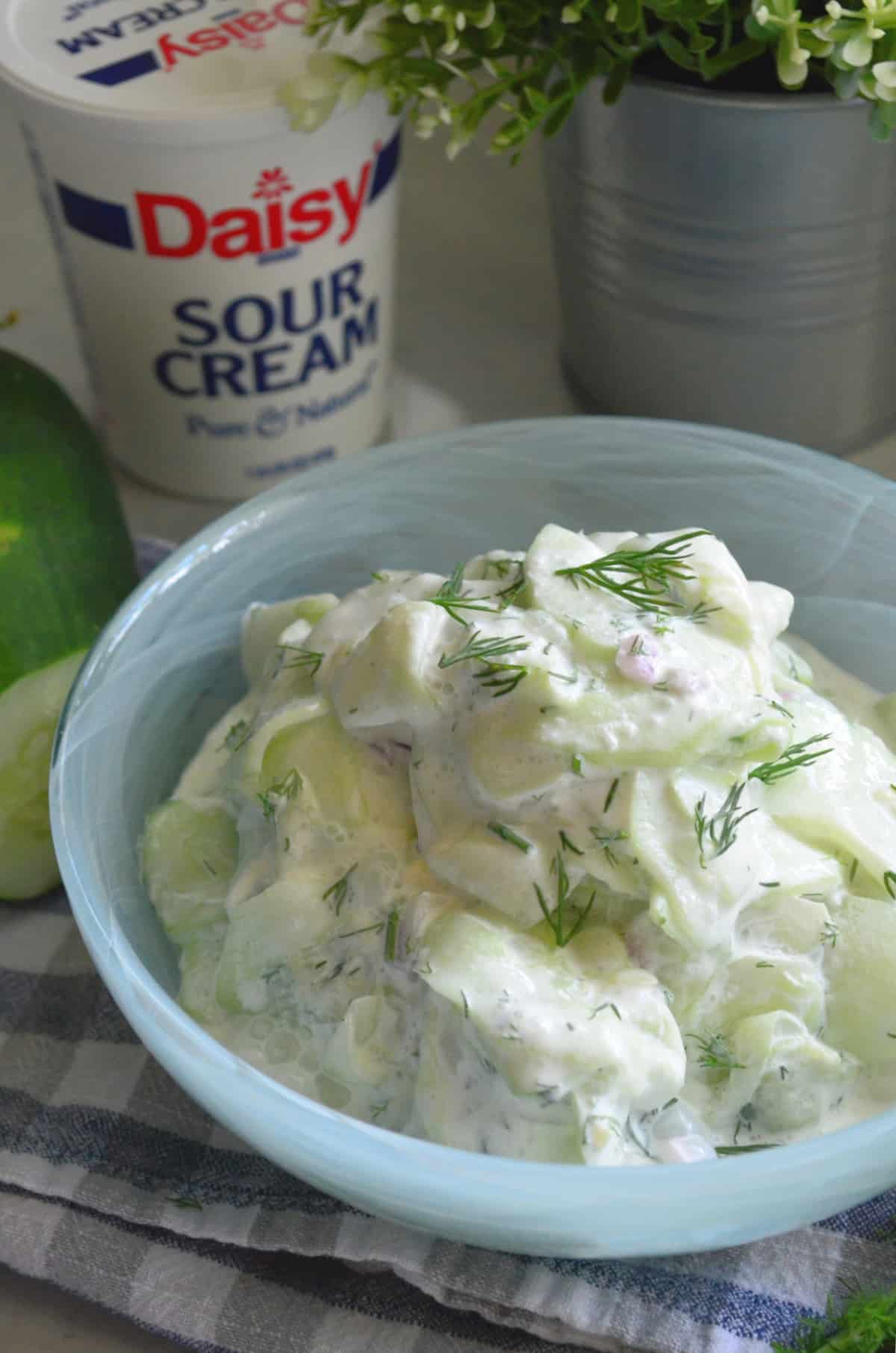 Close up of thinly sliced cucumbers with sour cream, fresh dill and red onion in blue bowl.