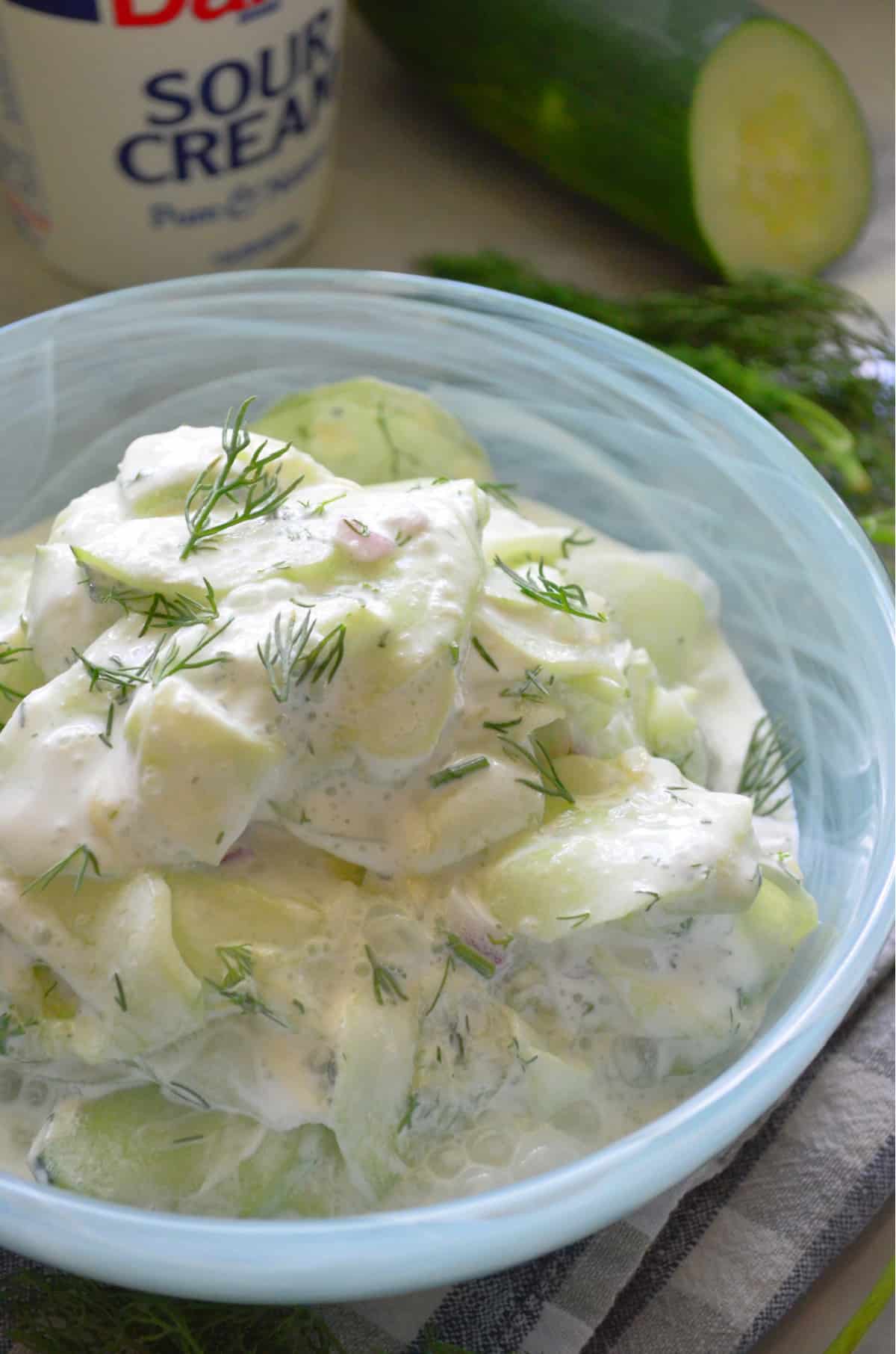 Up close of creamy thinly sliced cucumbers with dill in a blue bowl. 