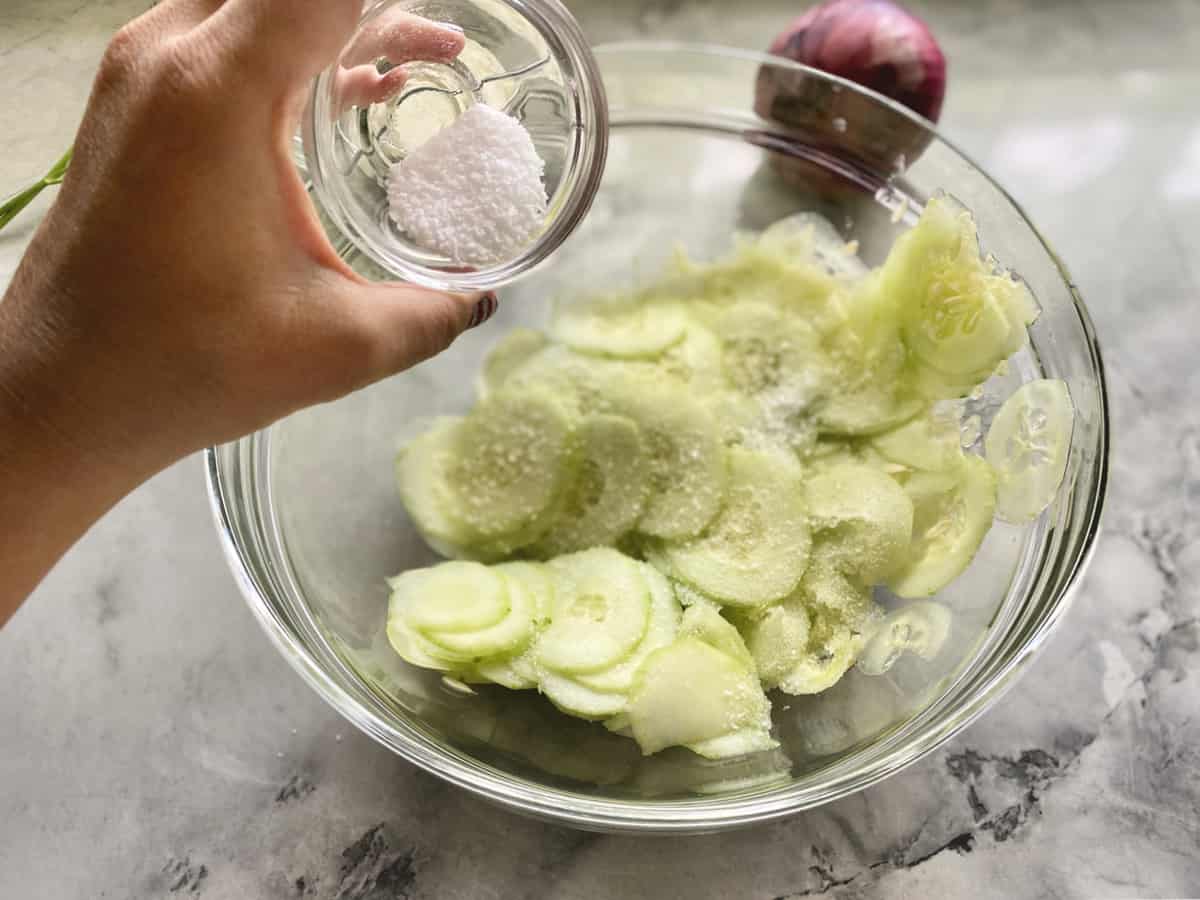 Hand pouring salt into a glass bowl of thinly sliced cucumbers on marble countertop.