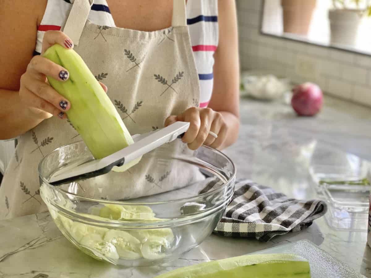Woman slicing cucumber using a hand-held mandolin slicer into a glass bowl.