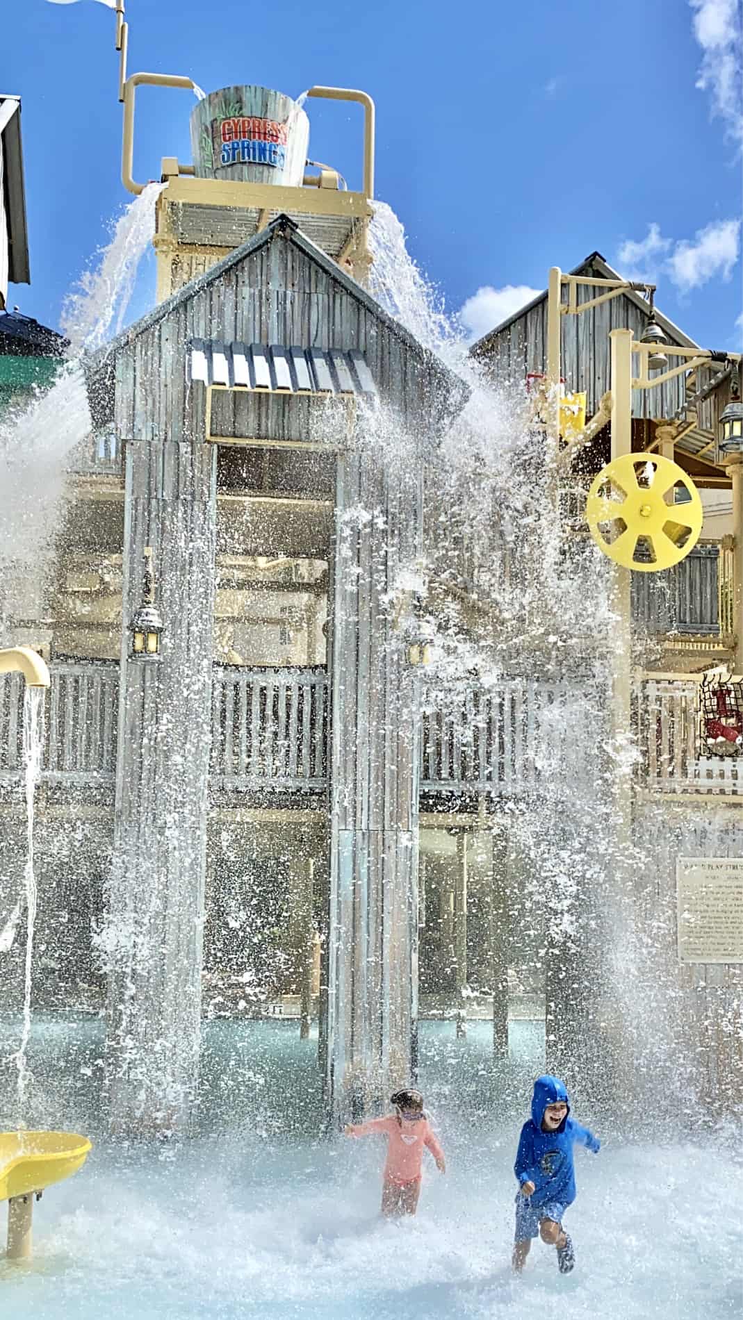 Two children; boy and girl, running at a splash park with water dumping on them. 