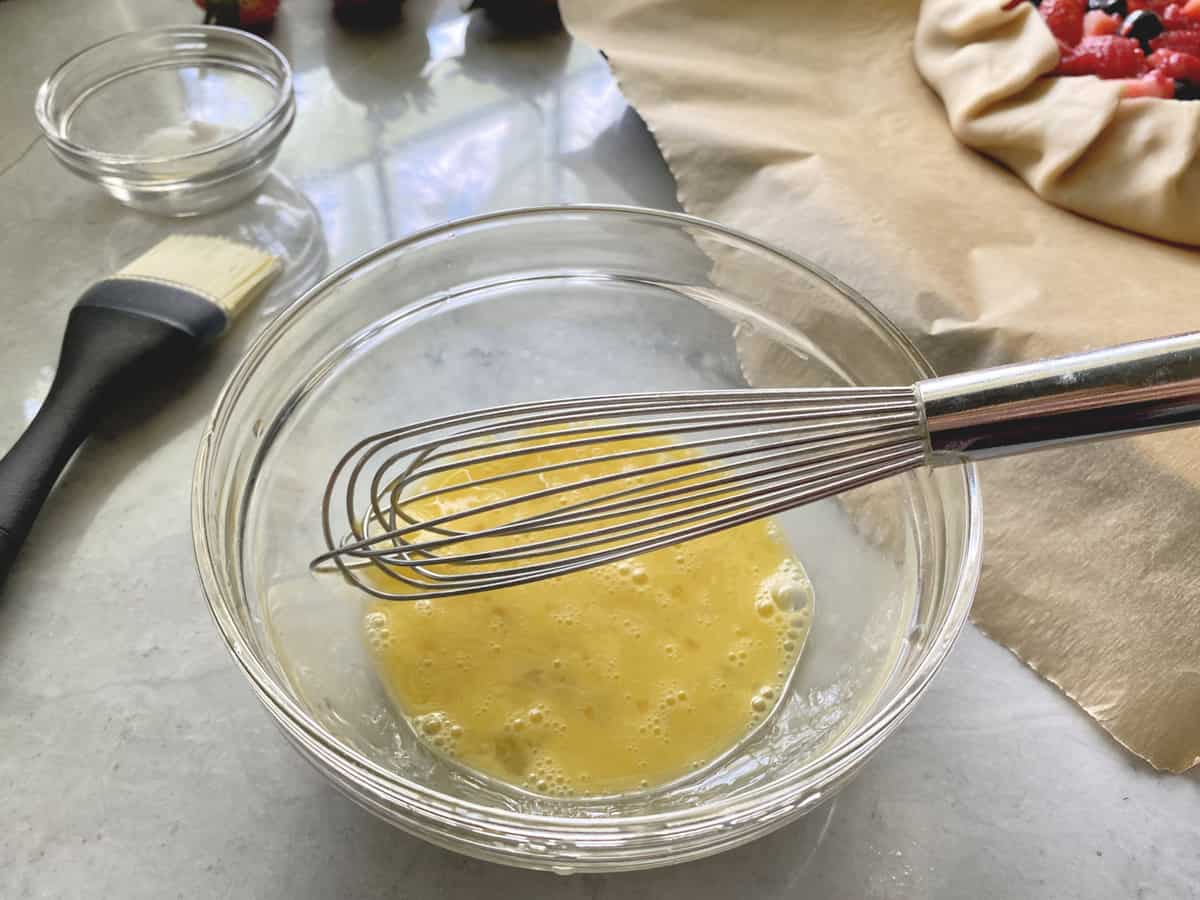 Glass bowl on a marble countertop with a whisk and scrambled egg in it. 