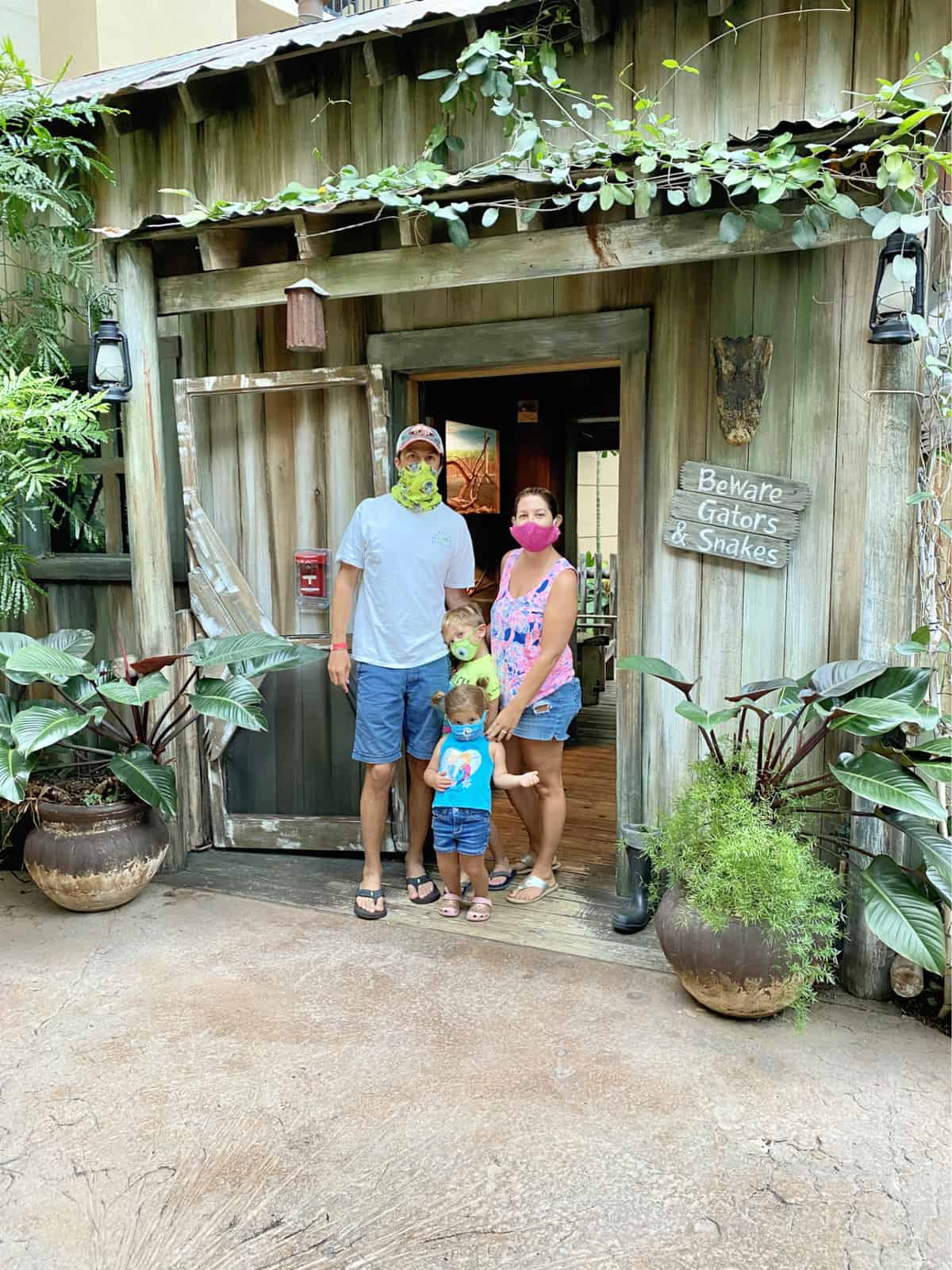 Family of four wearing face mask, standing in front of a cabin with greenery around them.