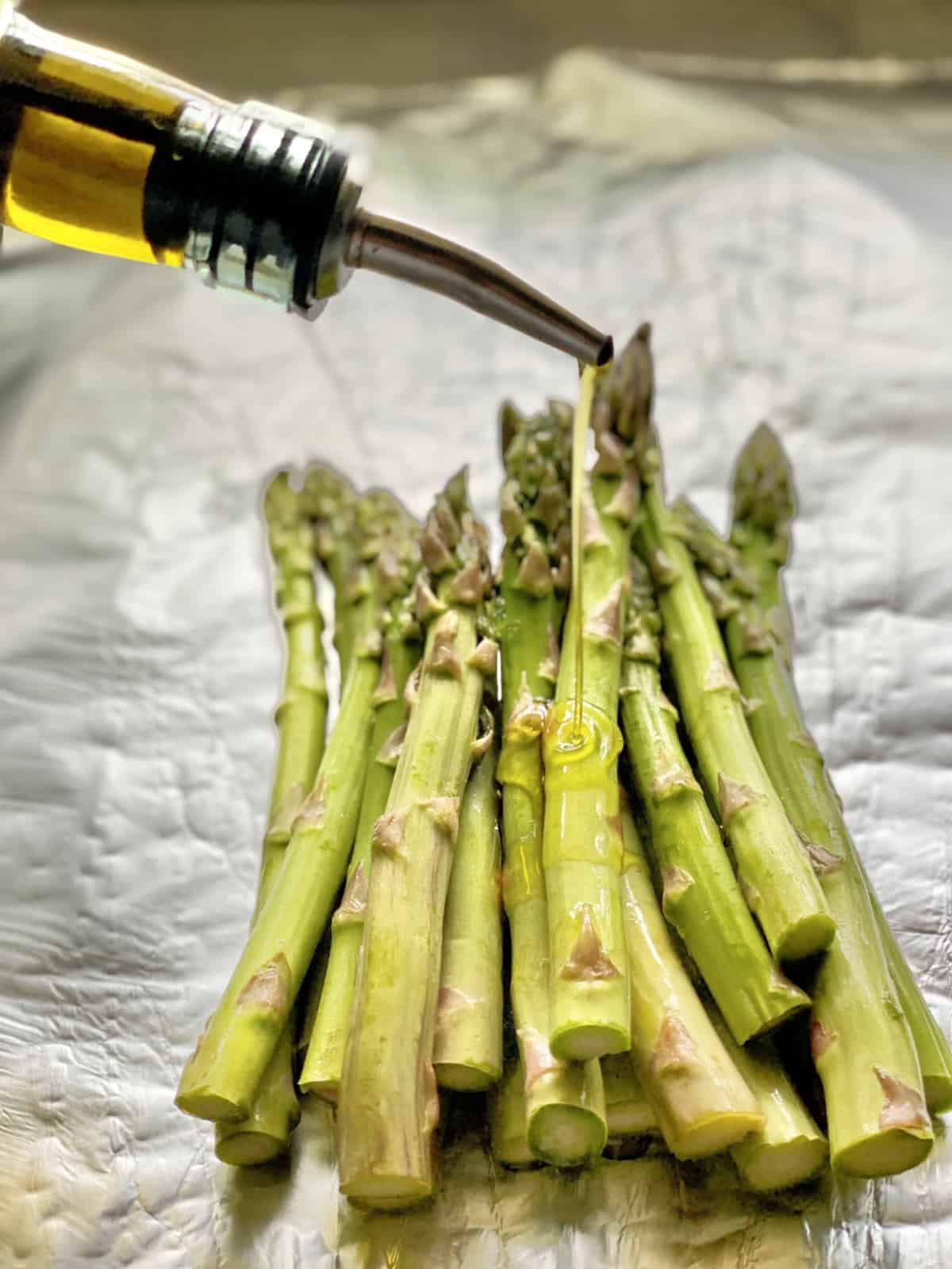 Bottle pouring oil on top of cut asparagus spears resting on foil.