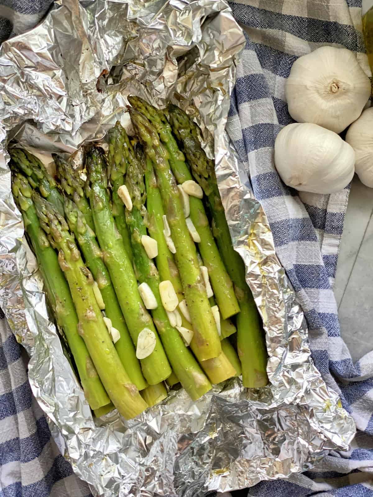 Top view of cooked asparagus with sliced garlic in foil pouch next to garlic bulbs.