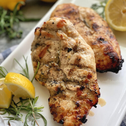 Grilled chicken breast on a white platter with fresh lemon wedges and rosemary sprigs.