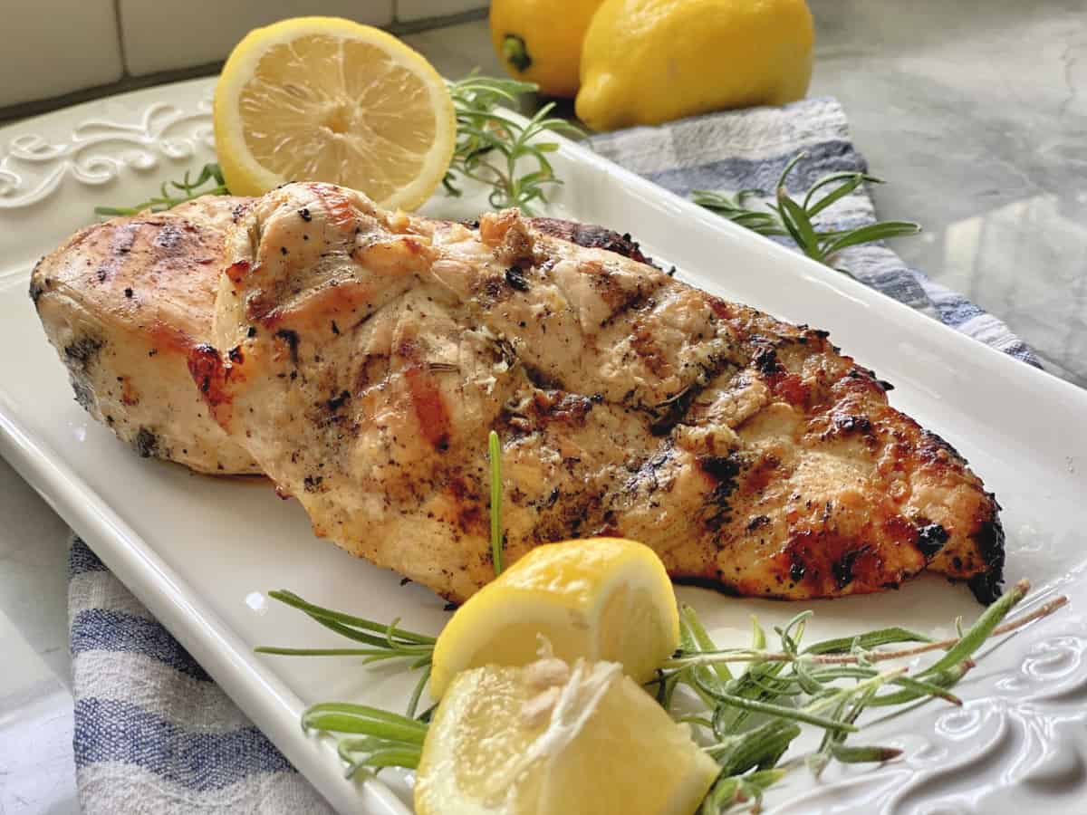 Grilled chicken breast on a white platter with lemon wedges and rosemary sprigs.