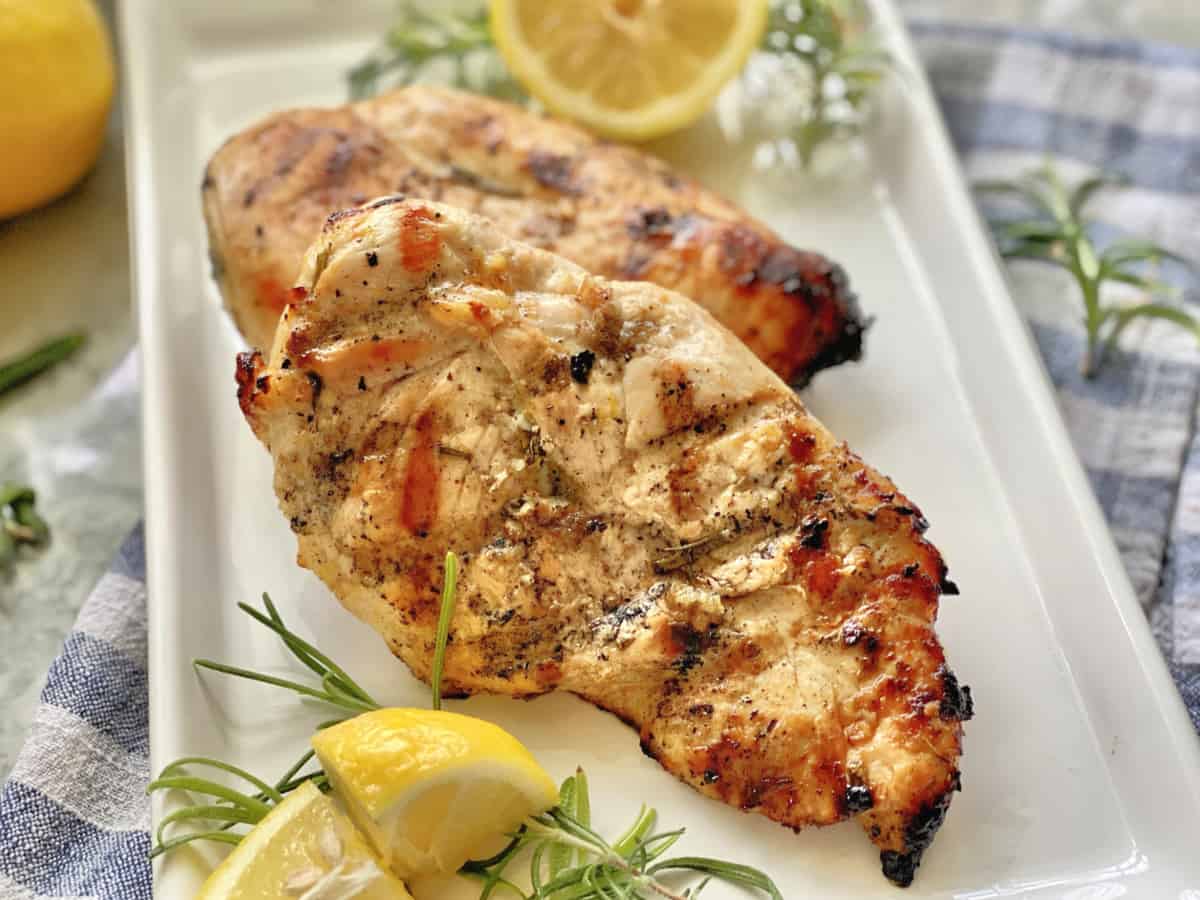 Close up of grilled chicken breast on white plate with fresh rosemary and lemon.