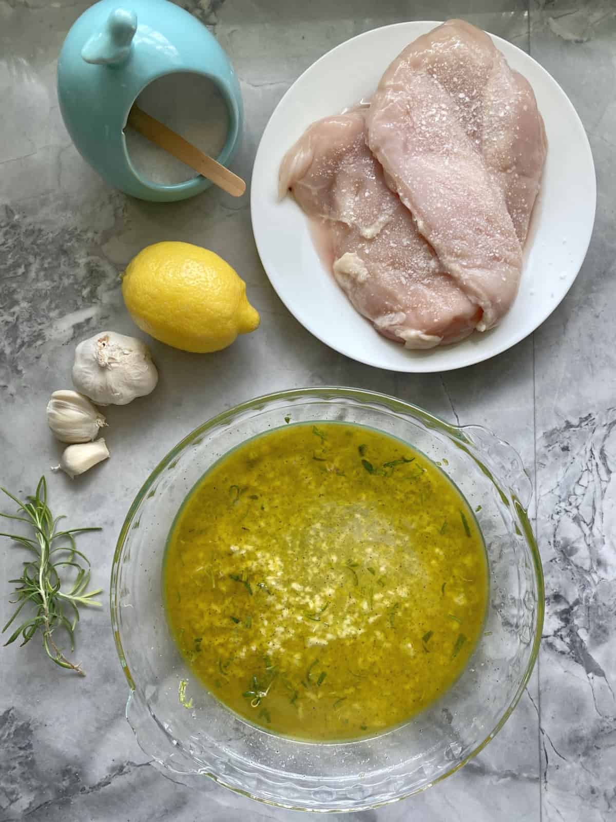 Salted raw chicken breast on a plate with lemon, garlic and a glass dish next to it.