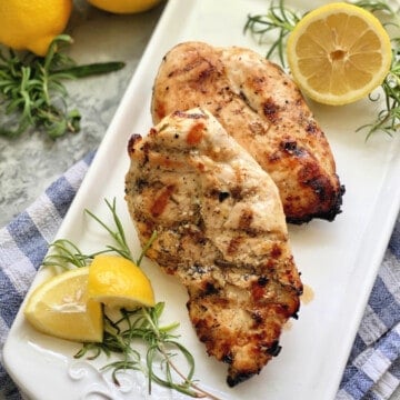 Two grilled chicken breast with lemon wedges and rosemary on a white platter.