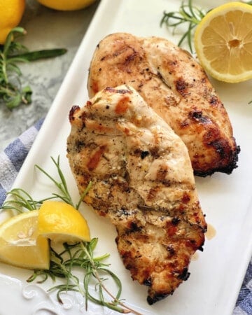 Two grilled chicken breast with lemon wedges and rosemary on a white platter.
