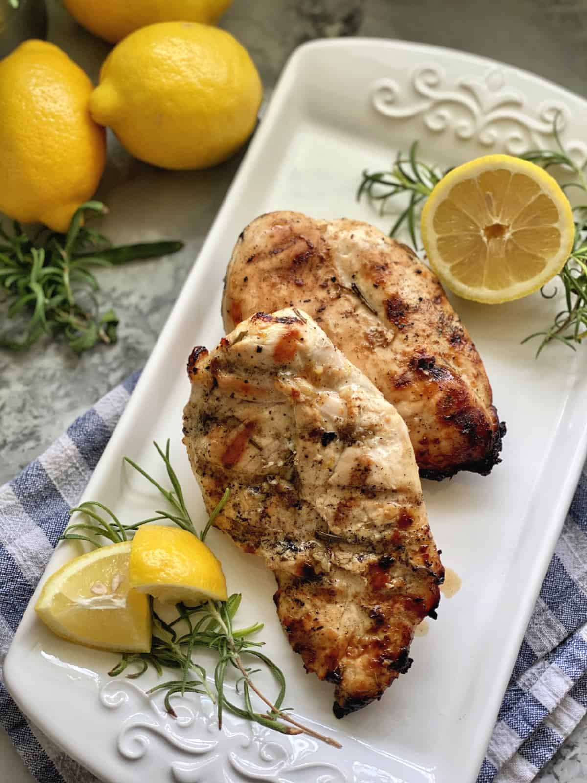 Top view of grilled chicken breast on a white platter with fresh lemon and rosemary.