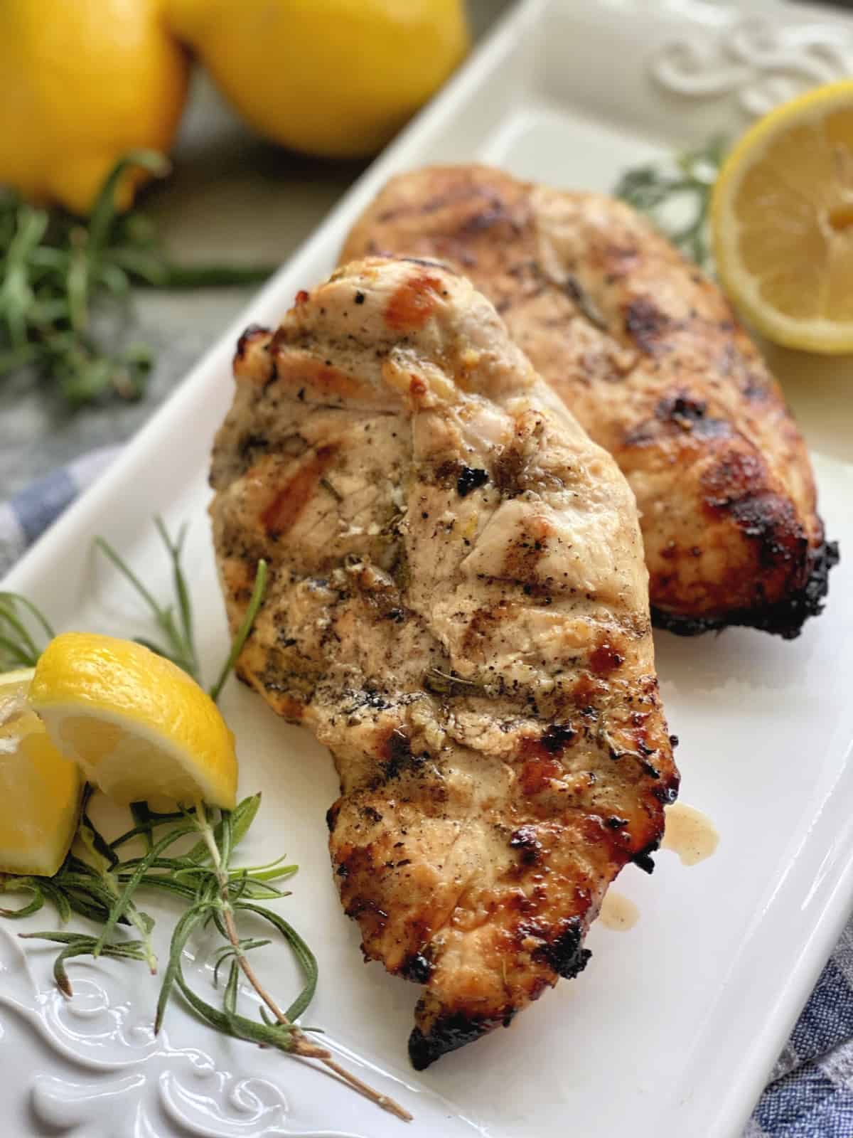 Grilled chicken breast on a white platter with fresh lemon wedges and rosemary sprigs.